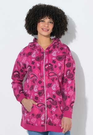 Vrouw met krullend haar, gekleed in een felroze hoodie met ritssluiting en zwart-witte paisley patronen, staand tegen een effen lichte achtergrond.
