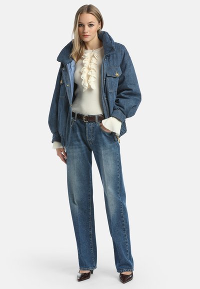 Chaqueta de mezclilla con un cuello alto, acentos en azul claro, combinada con un suéter blanco con volantes y pantalones de mezclilla de pierna ancha en azul claro. Detalles de cinturón marrón.