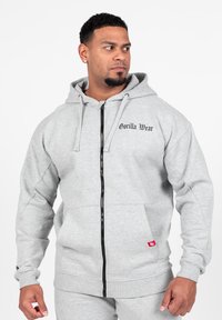 Grijze fleece hoodie met rits, voorzien van een logo aan de voorkant, zakken en een zwarte rits, met ribbels aan de mouwen en de tailleband.