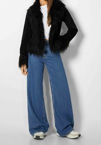 Giacca corta in pelliccia sintetica nera con maniche lunghe, abbinata a jeans blu in denim a vita alta e gamba larga, insieme a sneakers di colore chiaro.