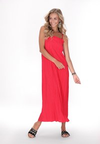 Maxi dress rosso plissettato con spalline sottili, caratterizzato da un colletto arricciato e un design fluido. Sandali neri aperti con un motivo intrecciato.