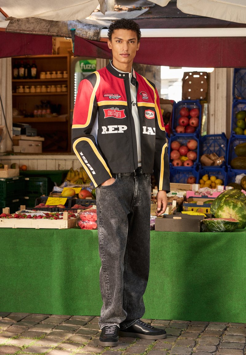 Jachetă scurtă neagră și roșie cu logo-uri, tricou gri, blugi negri și pantofi negri. Fundal de piață cu fructe și legume colorate.