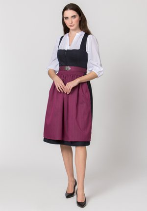 Frau stehend in traditionellem schwarzen Kleid mit weißer Bluse und violettem Schürze, Hände gefaltet, vor einfachem hellem Hintergrund.