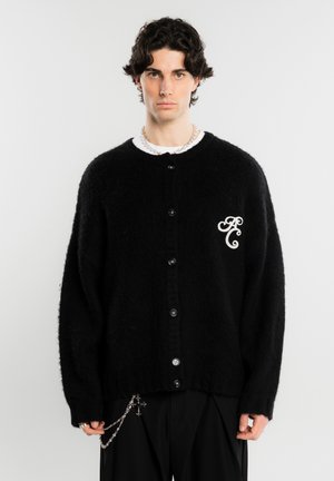 Jeune homme aux cheveux bouclés foncés porte un cardigan noir boutonné avec un logo blanc brodé et des accessoires en chaîne argentés sur un fond uni.