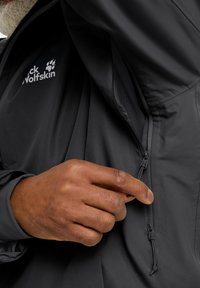 Giacca nera impermeabile con una texture liscia, dotata di chiusura a zip e linguette di sollevamento. Il logo "Jack Wolfskin" è esposto in modo prominente.