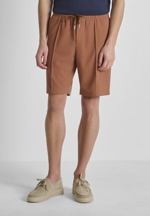 Uomo che indossa pantaloncini marroni con coulisse, maglietta blu navy e scarpe casual beige, in piedi contro uno sfondo chiaro e semplice.