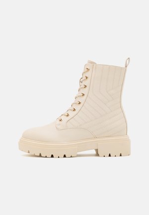 Botte de combat crème à lacets avec œillets dorés, panneaux latéraux matelassés, semelle épaisse et languette arrière sur fond blanc.