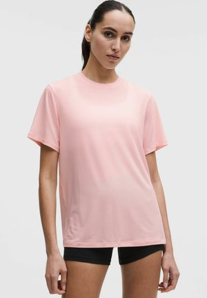 Kvinde med mørkt hår samlet i en hestehale, iført en løs, lyserød kortærmet skjorte og sorte shorts foran en ensfarvet, lys baggrund.