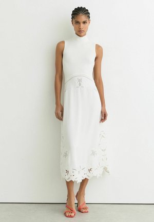 Fonott hajú nő, fehér, ujjatlan midi ruhában, virágmintás kivágásokkal, piros pántos magas sarkú szandálban, fehér fal előtt állva.