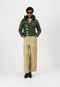 Groene glanzende pufferjack met een capuchon, ritssluiting en deurstiksel, gecombineerd met beige cargo broeken en zwarte enkellaarsjes.