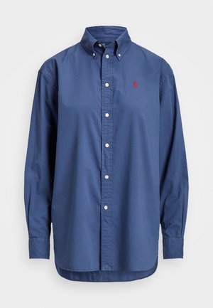 Chemise à manches longues bleue à boutons avec col et boutons blancs, arborant un petit logo rouge brodé sur la poitrine gauche.