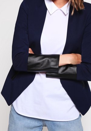Personne portant un blazer bleu marine avec des poignets de manches en cuir noir sur une chemise blanche à col et un jean bleu clair, bras croisés.