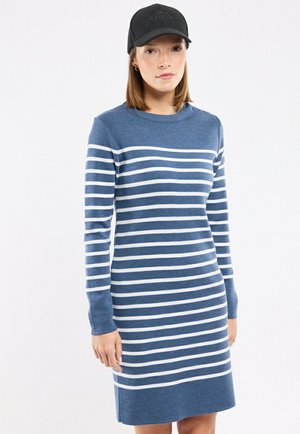 Femme portant une robe à manches longues à rayures horizontales bleues et blanches et une casquette de baseball noire, debout sur un fond blanc uni.