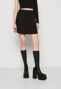 Mini-jupe noire en coton avec une taille cintrée, associée à des bottes en cuir noir montant jusqu'au genou, à talons et plateforme épaisse, avec une texture lisse.