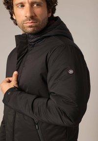 Veste noire isolante pour homme avec un col haut, une capuche ajustable et des poches zippées. Présente un logo sur la manche gauche. Texture lisse.