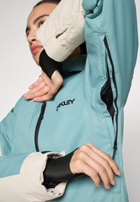 Turquoise and beige jacket with black cuffs; features zipper and mesh vent detail. Logo displayed on chest area.