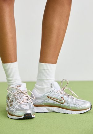 Beine mit weißen Crew-Socken und silbernen Nike Laufschuhen mit bronzefarbenen Akzenten, die auf einem grünen Teppich vor einem einfarbigen Hintergrund stehen.