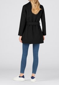 Cappotto nero con cintura, maniche lunghe e vestibilità aderente, abbinato a jeans skinny blu e sneakers bianche con accenti blu navy.