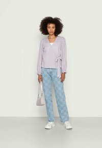 Top envolvente em gingham roxo com mangas bufantes longas, combinado com jeans quadriculados em azul claro. Acessórios incluem uma pequena bolsa lavanda e tênis brancos.