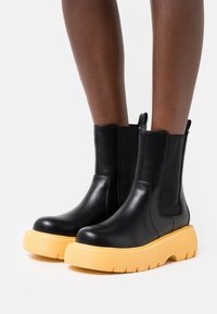 Botas de cuero negras hasta el tobillo con gruesas suelas amarillas, usadas sobre piernas de piel oscura, contra un fondo blanco liso.