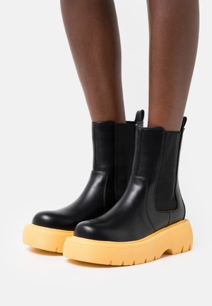 Botas de cuero negras hasta el tobillo con gruesas suelas amarillas, usadas sobre piernas de piel oscura, contra un fondo blanco liso.