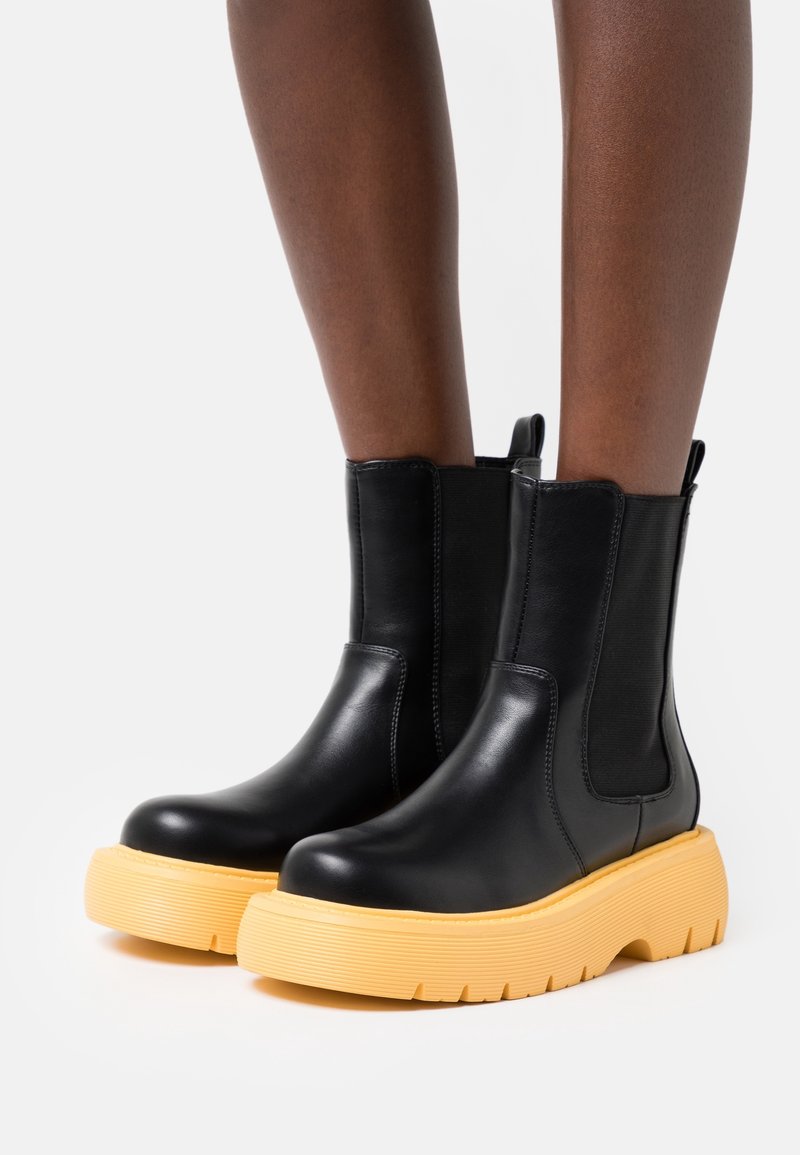 Botas de cuero negras hasta el tobillo con gruesas suelas amarillas, usadas sobre piernas de piel oscura, contra un fondo blanco liso.