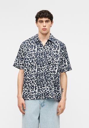 Nuori mies, jolla on päällään lyhythihainen, napillinen paita, jossa on mustavalkoinen abstrakti tekstiprintti, sekä vaaleansiniset farkut, seisoo yksinkertaisen taustan edessä.