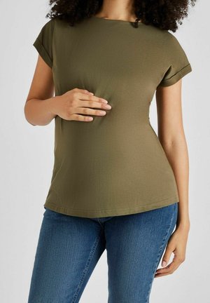 Kobieta w oliwkowej koszulce i niebieskich dżinsach, opierająca lewą rękę na brzuchu, a prawą rękę trzymającą obok siebie.