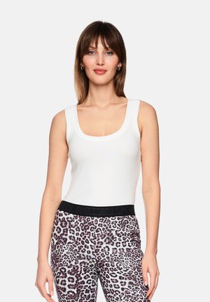 Frau trägt ein weißes ärmelloses Oberteil und Leggings mit Leopardenmuster und schwarzem Bund, steht vor einem schlichten hellen Hintergrund.