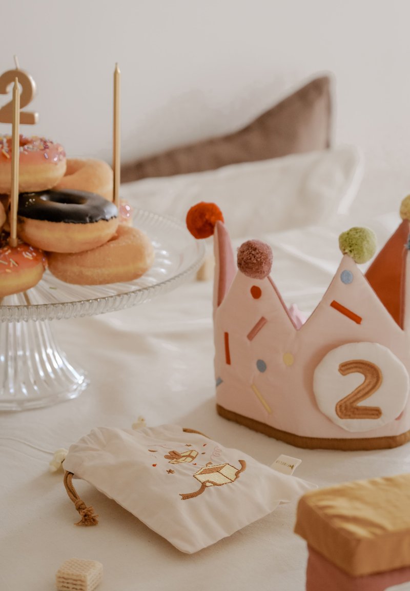 Rózsaszín korona színes pomponokkal, tetején a "2" szám; fehér vászonzacskó hímzéssel; vegyes fánkok üvegtálcán.