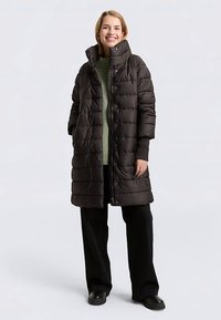 Lange, donkerbruine donsjas met een hoge kraag, doorgestikte textuur en knoopsluiting. Draagt over een groene trui met brede mouwen.