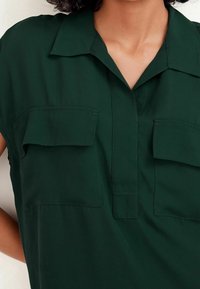 Blouse verte à manches courtes avec un col chemise, deux poches avant et une texture lisse et mate. Des détails de coutures soignées sont visibles.