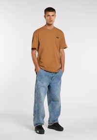 Brun bomulds t-shirt med broderet logo, parret med oversized blå denim jeans og sorte sneakers, stående foran en neutral baggrund.