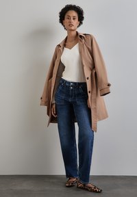 Beige Trenchcoat, weißes geripptes Oberteil, dunkelblaue Jeans mit ausgestellten Beinen und Ballerinas im Leopardenmuster; das Model steht vor einem neutralen Hintergrund.