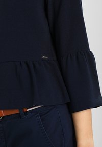 Marinblå peplumtop i texturerad tyg med utsvängd ärm och en liten logodetalj nära fållen. Kombinerad med marinblå byxor och ett brunt bälte.