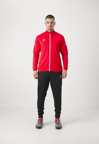 Sarkans sporta jaka ar pilnu rāvējslēdzēju un augstu apkakli, saskaņota ar melnajiem treniņbiksēm; pelēki sporta apavi ar sarkaniem akcentiem.