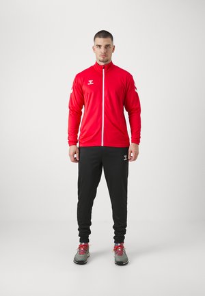 Giacca sportiva rossa con zip completa e colletto alto, abbinata a pantaloni tuta neri; scarpe da ginnastica grigie con dettagli rossi.