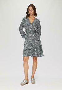 Robe portefeuille en tissu bleu foncé, avec des manches longues, une taille à nouer et un ourlet évasé. Associée à des sabots en métal argenté.
