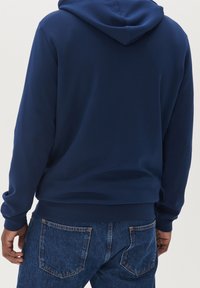 Felpa blu navy con una consistenza morbida, polsini e orlo a coste. Presenta un cappuccio con cordini e una vestibilità comoda, abbinata a jeans in denim.