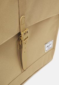 Tan canvas backpack with a textured surface, featuring a plastic buckle closure and a white brand label on the side.