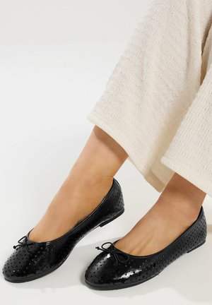 Stopy w czarnych ażurowych balerinach z małymi kokardkami, w połączeniu z luźnymi, teksturowanymi, kremowymi spodniami.