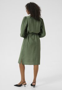Robe verte à manches bouffantes, fabriquée en tissu léger. Elle dispose d'une fente sur le côté et d'une ceinture assortie. Des escarpins noirs à brides arrières complètent le look.