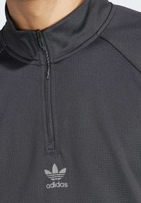Czarna sportowa bluza z półzamkniętym kołnierzem, teksturowaną tkaniną i srebrnym logo Adidasa na piersi. Posiada raglanowe rękawy.