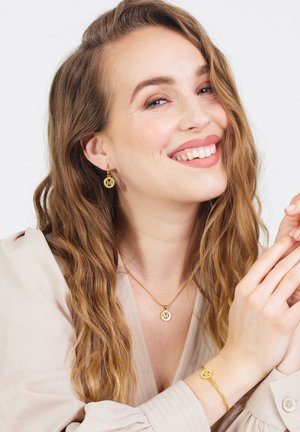 Gouden sieradenset met een cirkelvormige hangerketting, bijpassende oorbellen en armband. Het ontwerp omvat een logodetail en een gladde textuur.