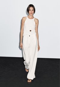 Witte mouwloze top gecombineerd met losse, witte broeken. De outfit heeft een eenvoudig ontwerp en een lange ketting met een ronde hanger.