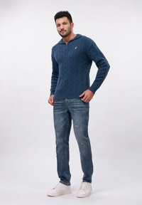 Gestrickter marineblauer Pullover mit einem Reißverschlusskragen und einem Zopfmuster. Kombiniert mit zerrissenen blauen Jeans und weißen Sneakern.