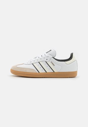 Hvid Adidas Samba-sneaker med sorte striber, beige ruskindstå, gummisål og teksten "Samba" på siden, vist fra den indvendige side.