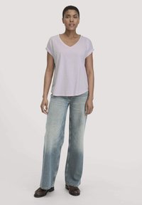 Vrouw staat met een lichtpaarse V-hals korte mouw shirt, wijdvallende vervaagde blauwe jeans, en donkerbruine schoenen tegen een effen achtergrond.