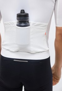 Άσπρο αθλητικό μπλουζάκι με τρεις πίσω τσέπες, που κρατάει ένα μαύρο παγούρι νερού, φορεμένο από ένα άτομο με μαύρο ποδηλατικό σορτς.