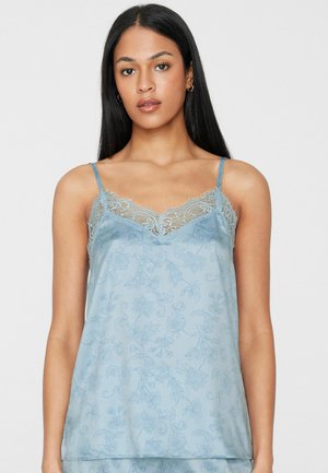 Vrouw met lang donker haar, gekleed in een lichtblauwe bloemenprint cami met kanten rand en dunne schouderbandjes, kijkend naar voren tegen een effen achtergrond.